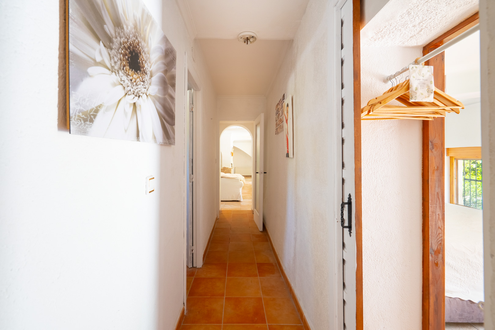 Luminoso pasillo con suelos de baldosas terracota y paredes blancas texturizadas. Se aprecian cuadros decorativos y un acceso a una sala con arco, además de un espacio con perchas de madera. Luminoso pasillo con suelos de baldosas terracota y paredes blancas texturizadas. Se aprecian cuadros decorativos y un acceso a una sala con arco, además de un espacio con perchas de madera.