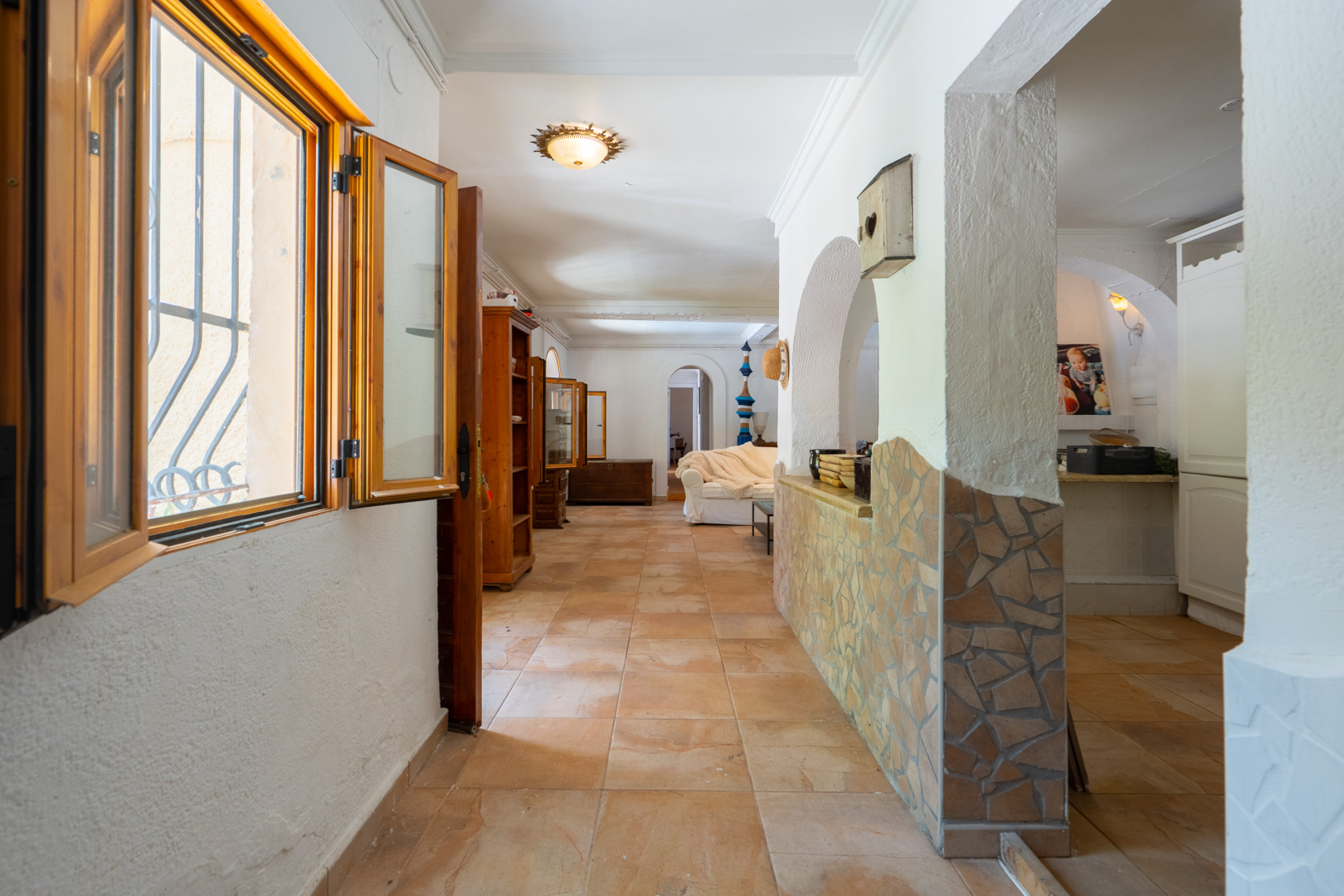 Pasillo luminoso con ventana de madera y rejas, suelo de baldosas de terracota y una pared baja con mosaico. Conduce a una sala de estar con arcos y muebles de madera. Pasillo luminoso con ventana de madera y rejas, suelo de baldosas de terracota y una pared baja con mosaico. Conduce a una sala de estar con arcos y muebles de madera.