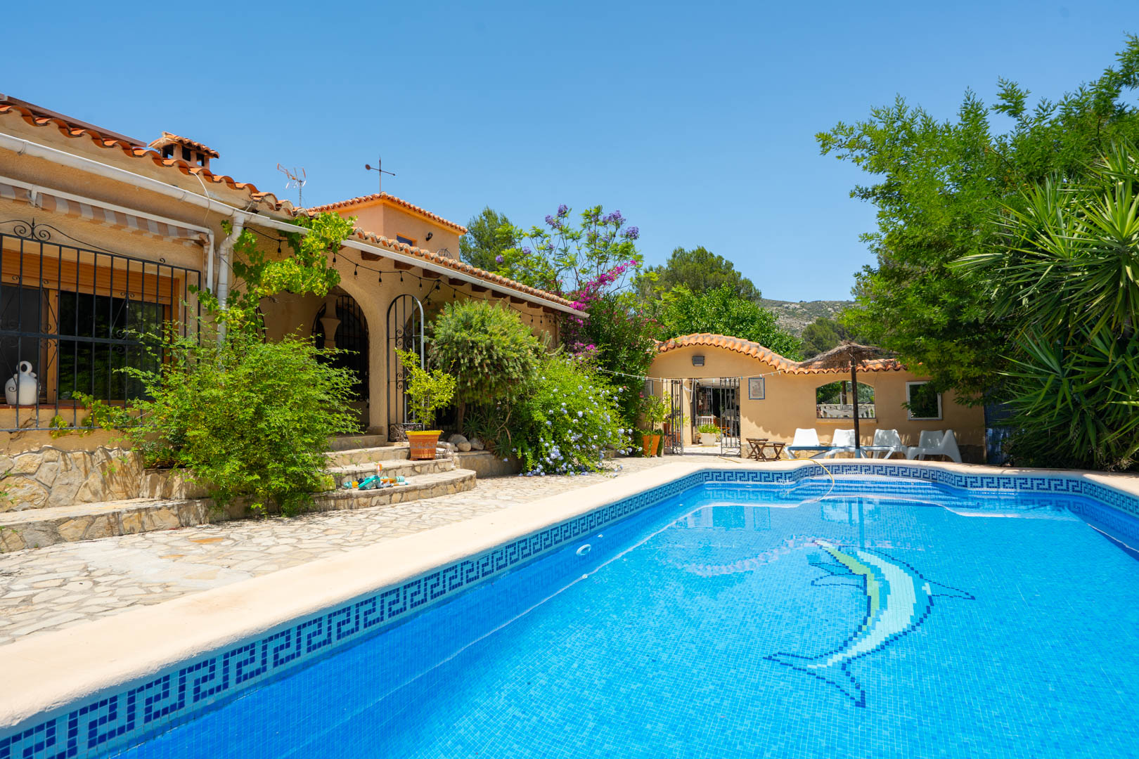 Piscina de mosaico azul con diseño de delfín y borde de greca, rodeada por un patio de piedra y edificios de estilo mediterráneo con abundante vegetación bajo un cielo despejado. Piscina de mosaico azul con diseño de delfín y borde de greca, rodeada por un patio de piedra y edificios de estilo mediterráneo con abundante vegetación bajo un cielo despejado.