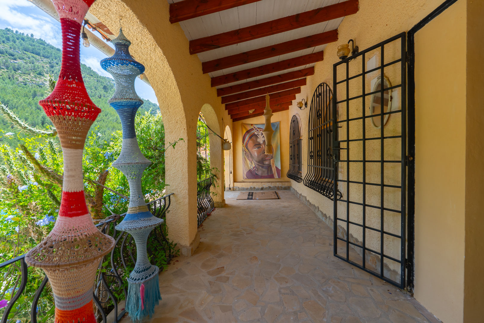 Terraza cubierta con suelo de piedra irregular, vigas de madera vistas y arcos que enmarcan la vista al valle. Decorada con lámparas de ganchillo y arte mural. Terraza cubierta con suelo de piedra irregular, vigas de madera vistas y arcos que enmarcan la vista al valle. Decorada con lámparas de ganchillo y arte mural.