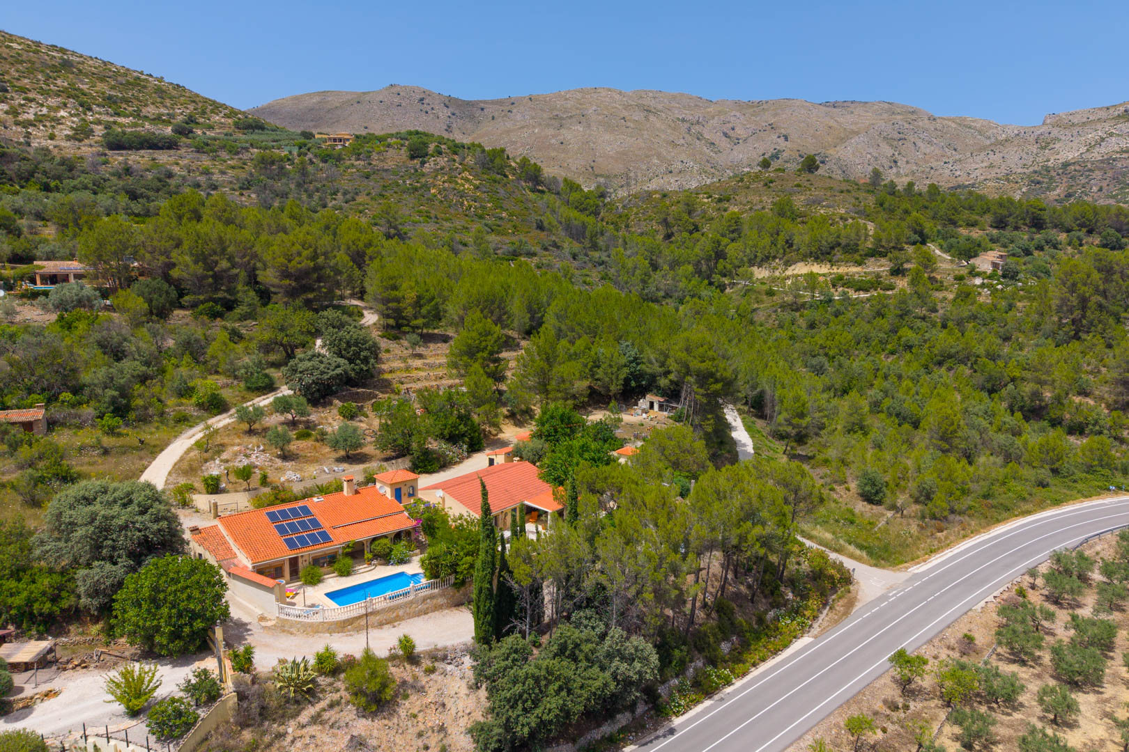 Vista aérea de la finca con dos viviendas, piscina y paneles solares, en un entorno montañoso y boscoso con acceso por carretera. Vista aérea de la finca con dos viviendas, piscina y paneles solares, en un entorno montañoso y boscoso con acceso por carretera.
