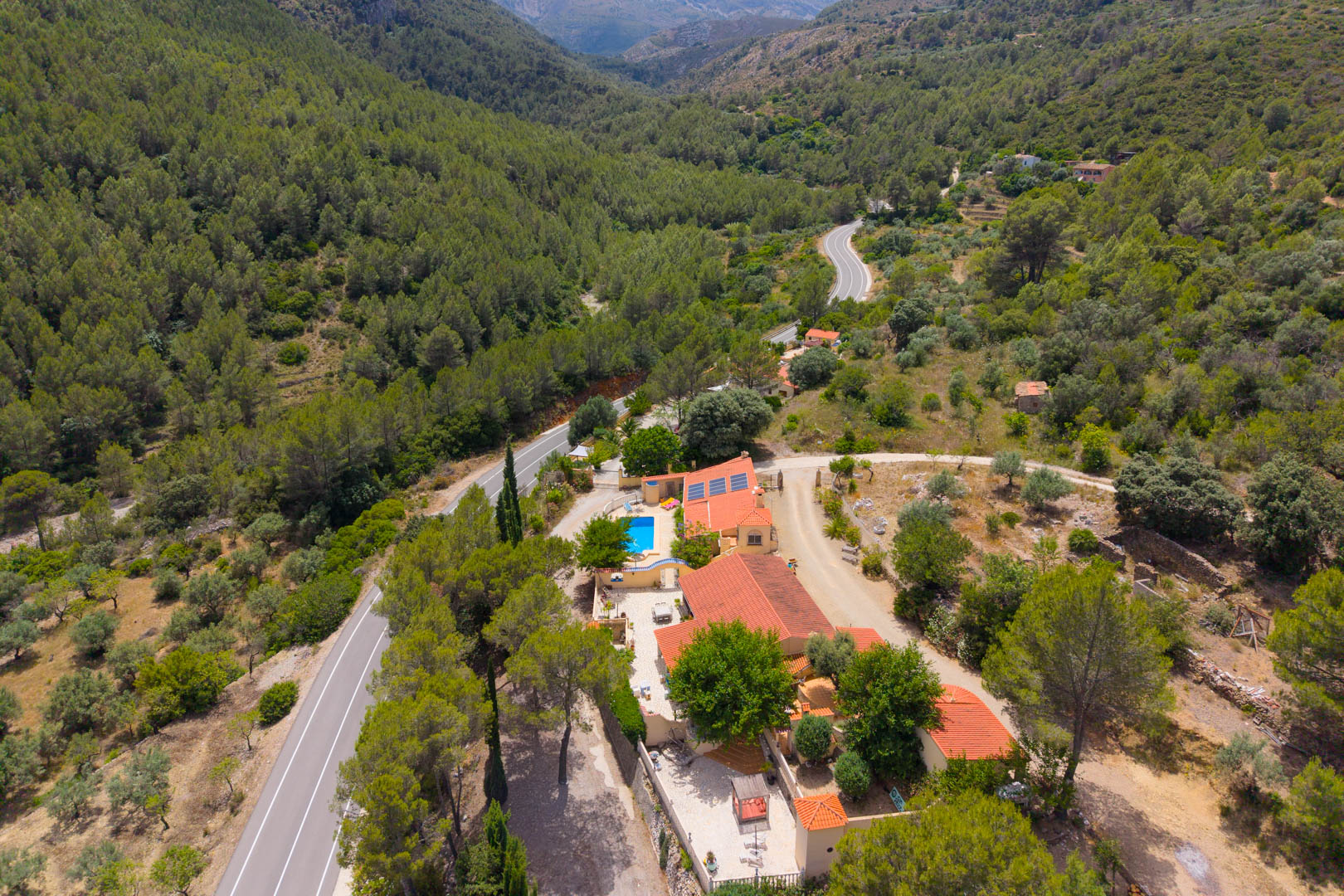 Vista aérea de la finca con dos viviendas, techos de teja y piscina azul, enclavada entre densos bosques verdes y una carretera sinuosa en un valle montañoso. Paneles solares visibles. Vista aérea de la finca con dos viviendas, techos de teja y piscina azul, enclavada entre densos bosques verdes y una carretera sinuosa en un valle montañoso. Paneles solares visibles.