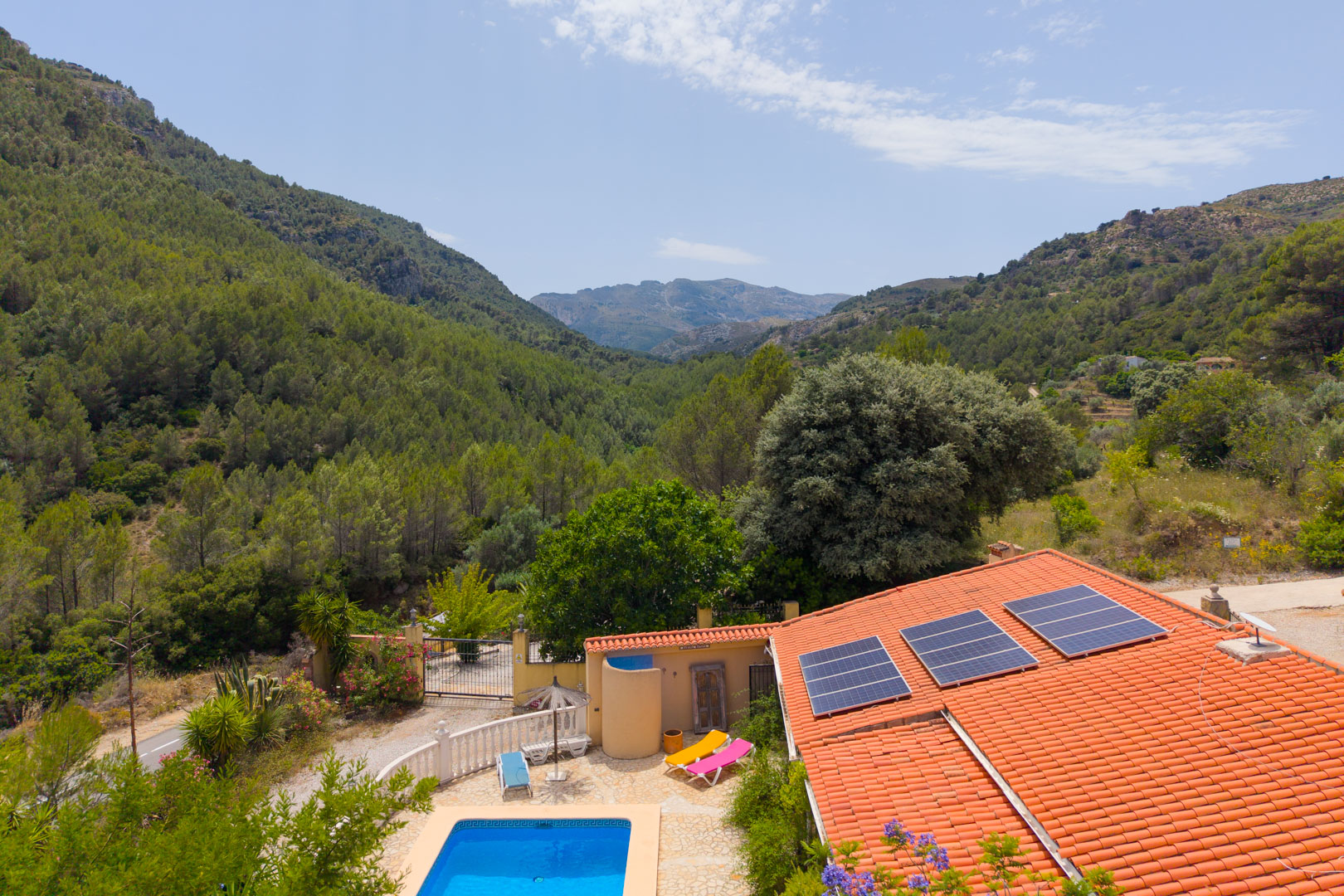Vista aérea de la finca con tejado de teja, paneles solares y piscina azul, enmarcada por montañas boscosas y vegetación mediterránea. Vista aérea de la finca con tejado de teja, paneles solares y piscina azul, enmarcada por montañas boscosas y vegetación mediterránea.