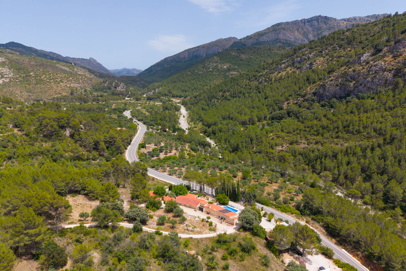 Vista aérea de la finca situada en un valle montañoso y boscoso, con piscina, paneles solares y edificios de tejado terracota, junto a una carretera serpenteante. Vista aérea de la finca situada en un valle montañoso y boscoso, con piscina, paneles solares y edificios de tejado terracota, junto a una carretera serpenteante.