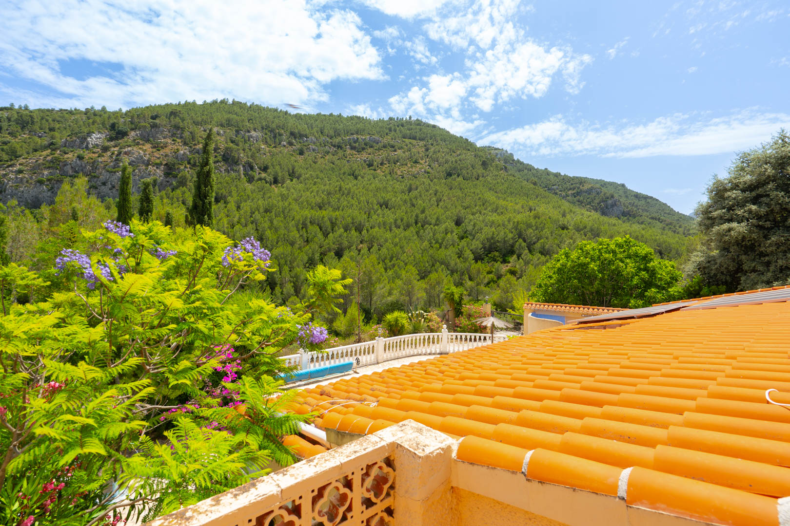 Vista desde una terraza con tejas de barro naranjas, una balaustrada decorativa y flores de jacarandá, enmarcada por montañas boscosas. Vista desde una terraza con tejas de barro naranjas, una balaustrada decorativa y flores de jacarandá, enmarcada por montañas boscosas.