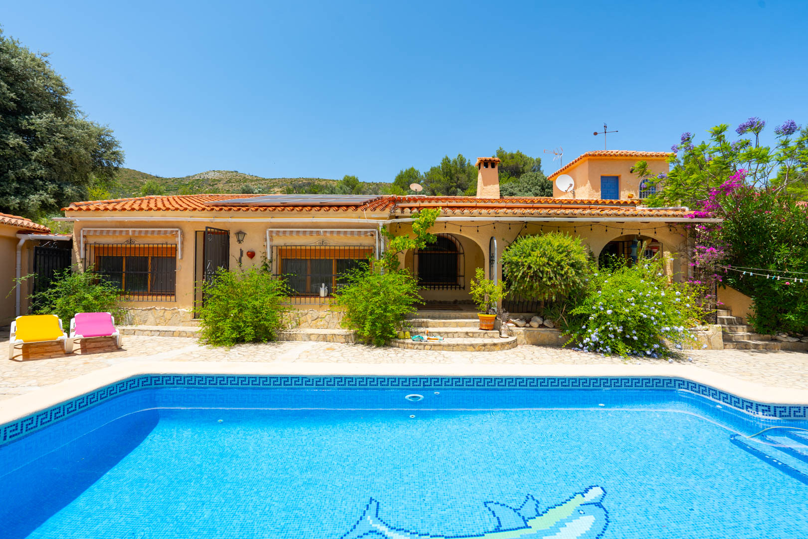 Vista exterior de las dos viviendas con tejados de teja, piscina de mosaico azul con delfín y paneles solares. Rodeada de exuberante vegetación mediterránea y montañas. Vista exterior de las dos viviendas con tejados de teja, piscina de mosaico azul con delfín y paneles solares. Rodeada de exuberante vegetación mediterránea y montañas.