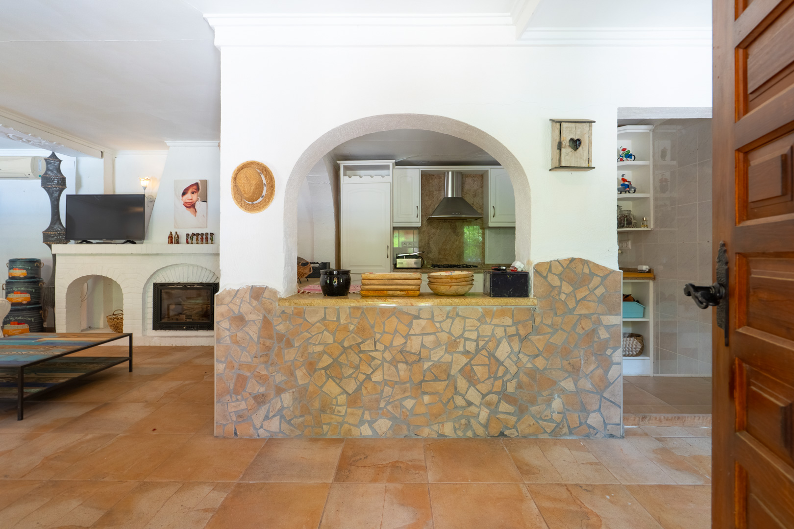 Zona de estar de planta abierta con suelos de terracota, chimenea blanca y paso arqueado con media pared de mosaico de piedra hacia la cocina semiabierta. Zona de estar de planta abierta con suelos de terracota, chimenea blanca y paso arqueado con media pared de mosaico de piedra hacia la cocina semiabierta.