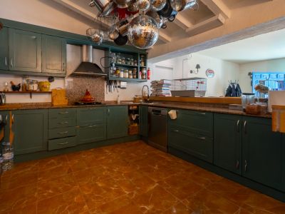 Amplia cocina con muebles en verde oscuro, encimeras de granito y suelo de baldosas de terracota. Su diseño semiabierto conecta con una estancia adyacente, destacando vigas vistas y decoración ecléctica.