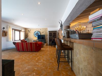 Amplia sala de estar con barra rústica revestida de piedra y tres taburetes, conectando con un salón que tiene sofá rojo, suelo de baldosas y un gran reloj circular en la pared de piedra.