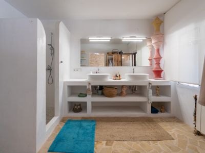 Amplio baño con doble lavabo integrado en encimera blanca, ducha de obra con pared curva y suelo de baldosas de piedra irregular. Espejo con iluminación.
