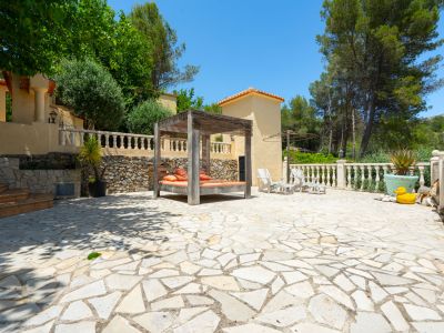 Amplio patio exterior con pavimento de losas irregulares de tonos claros, una cama balinesa de madera con cojines naranjas y sillas Adirondack blancas, rodeado de jardines aterrazados y árboles maduros.