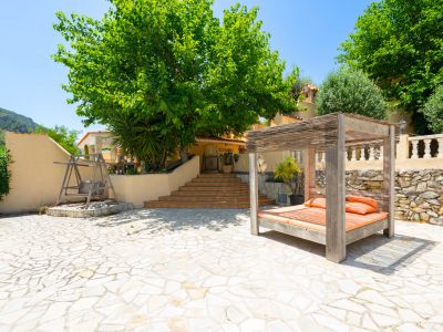 Amplio patio exterior con pavimento de piedra irregular, destacando una cama balinesa de madera con dosel de bambú y un banco columpio, con escaleras al edificio.