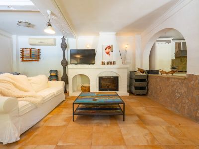Amplio salón con chimenea de ladrillo blanco y TV integrada, mesa de centro de madera multicolor única y suelo de baldosas de terracota. Un arco conecta con la cocina semiabierta.