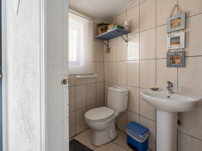 Baño auxiliar compacto con azulejos beige, inodoro y lavabo de pedestal blancos, luz natural de una ventana pequeña y estante decorativo.