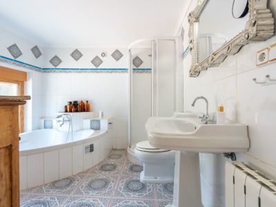 Baño con bañera esquinera, lavabo de pedestal y ducha. Presenta azulejos blancos con cenefas decorativas y suelo hidráulico de patrones geométricos.