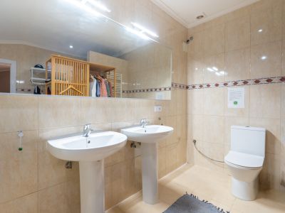 Baño con dos lavabos de pedestal, paredes alicatadas en tonos beige con cenefa decorativa y un amplio espejo que refleja un perchero de madera.