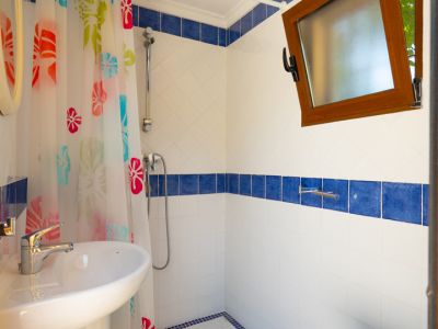 Baño con lavabo de pedestal blanco, ducha con cortina floral colorida y paredes de azulejos blancos con cenefa azul. Una ventana de madera abierta aporta luz natural.