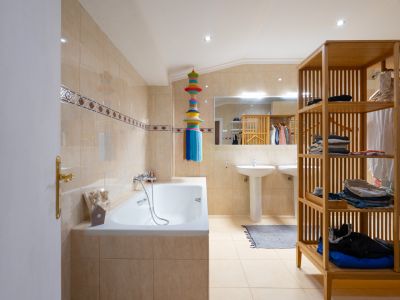 Baño con paredes de azulejos beige claro y cenefa decorativa, bañera y dos lavabos de pedestal. Un gran espejo refleja un mueble de estanterías de madera, iluminado por focos empotrados.