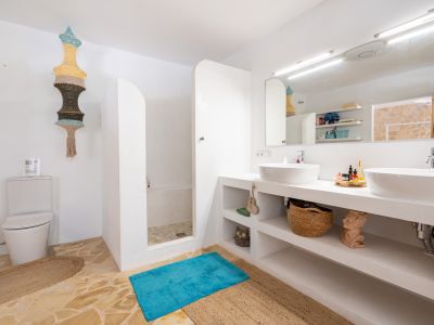 Baño luminoso con doble lavabo sobre encimera, ducha abierta con suelo de piedra irregular y paredes blancas. Destaca una lámpara colgante textil y el suelo de baldosas de piedra natural.
