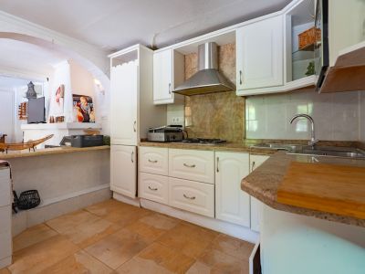 Cocina semiabierta con armarios blancos tradicionales, encimeras de granito y placa de gas. Dispone de fregadero doble y suelo de baldosas de terracota que conecta con una barra.
