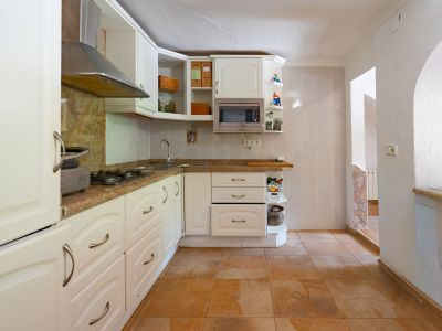 Cocina semiabierta con muebles blancos, encimeras de granito y electrodomésticos integrados. El suelo de baldosas terracota complementa las paredes claras y el arco de acceso.