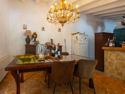 Comedor con mesa de madera oscura y cristal, sillas tapizadas marrones, y una lámpara de araña dorada. Conecta con una barra de cocina revestida de piedra y vigas vistas.