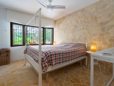 Dormitorio con cama con dosel, pared de acento y suelo de piedra irregular, y ventanas con rejas que ofrecen vistas a la vegetación exterior.