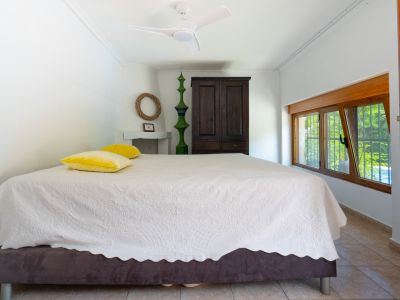 Dormitorio con cama de colcha blanca estampada, armario de madera oscura y ventana de madera que da a la vegetación. Incluye ventilador de techo y suelo de baldosas.