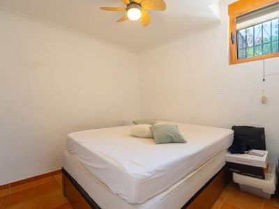 Dormitorio con cama doble, paredes blancas texturizadas y suelo de baldosas de terracota. Ventana de madera con rejas de seguridad y ventilador de techo.