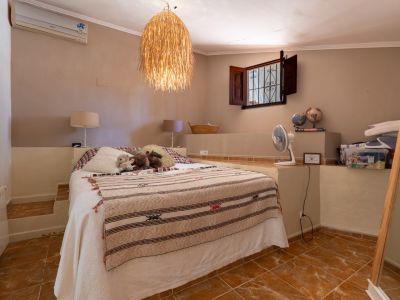 Dormitorio con cama doble sobre plataforma elevada, suelo de baldosas de terracota y lámpara colgante de mimbre. Ventana abierta con contraventanas de madera oscura y reja de hierro.