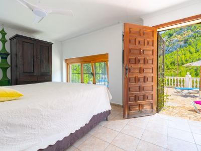 Dormitorio con suelo de baldosas claras, armario de madera oscura y ventilador de techo. Una puerta de madera abierta da a una terraza con vistas a la montaña y tumbonas.