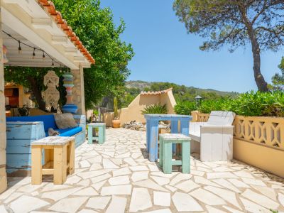 Encantador patio exterior con pavimento de piedra irregular, área de asientos cubierta con sofá azul y luces, junto a muebles de madera decapada y vegetación.