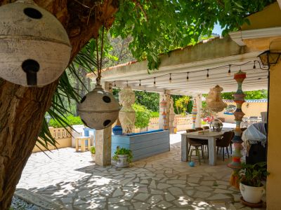 Encantadora zona de comedor exterior cubierta con mesa de madera rústica sobre pavimento de piedra, adornada con pilares artesanales y lámparas de cuentas, bajo un árbol frondoso con campanas.