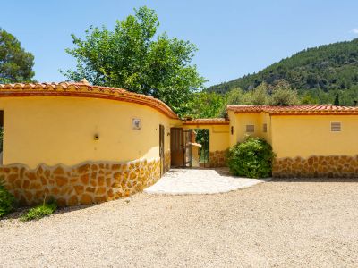 Entrada a la propiedad con camino de grava y sendero pavimentado, flanqueada por muros de estuco amarillo con zócalo de piedra natural y tejas de terracota, con fondo montañoso.