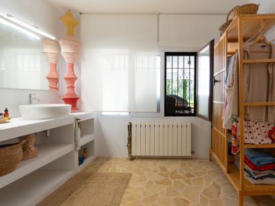 Espacio de baño funcional con lavabo sobre encimera blanca y estantes abiertos, sobre un suelo de baldosas de piedra irregular. Incluye un armario abierto de madera y ventana con reja de seguridad.