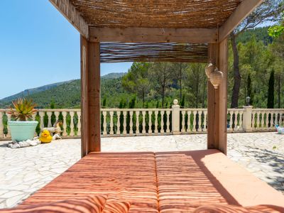 Espaciosa terraza exterior con pérgola de madera y techo de paja, cubriendo una doble tumbona. Ofrece vistas panorámicas a las montañas y está delimitada por una balaustrada clásica.