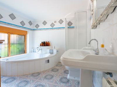 Este luminoso baño presenta una bañera esquinera alicatada bajo una ventana de madera, junto a un lavabo de pedestal y una cabina de ducha. Los azulejos estampados decoran el suelo y el borde de la pared.