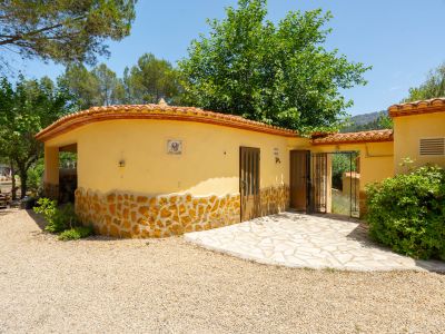 Exterior de una construcción auxiliar curva de color amarillo con base de piedra natural y tejado de tejas rojas. Incluye puerta de madera, azulejo decorativo y entrada pavimentada sobre grava.