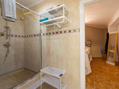 Funcional baño con ducha de azulejos beige, mampara de cristal y cenefa decorativa. Una puerta abierta revela un dormitorio adyacente con suelo de baldosas de terracota y espejo de cuerpo entero.
