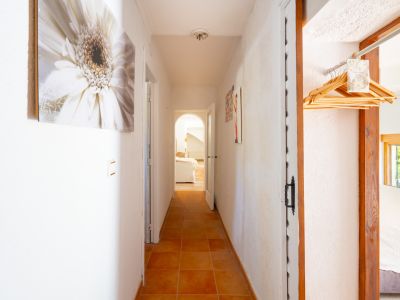 Luminoso pasillo con suelos de baldosas terracota y paredes blancas texturizadas. Se aprecian cuadros decorativos y un acceso a una sala con arco, además de un espacio con perchas de madera.