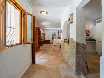 Pasillo luminoso con ventana de madera y rejas, suelo de baldosas de terracota y una pared baja con mosaico. Conduce a una sala de estar con arcos y muebles de madera.