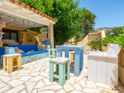 Terraza cubierta con suelo de losas irregulares, mobiliario rústico de madera pintada y coloridas lámparas colgantes, rodeada de vegetación.