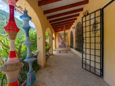 Terraza cubierta con suelo de piedra irregular, vigas de madera vistas y arcos que enmarcan la vista al valle. Decorada con lámparas de ganchillo y arte mural.