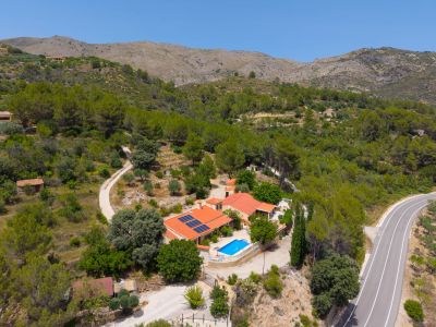 Vista aérea de la finca con dos edificios de tejado rojo, piscina azul y paneles solares, rodeada de bosque y montañas, con carretera serpenteante.