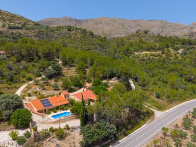 Vista aérea de la finca con dos viviendas, piscina y paneles solares, en un entorno montañoso y boscoso con acceso por carretera.