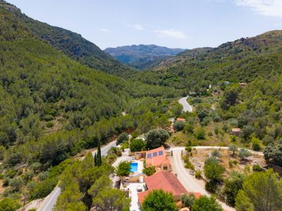 Vista aérea de la finca con dos viviendas, piscina y paneles solares, enclavada en un valle montañoso con densa vegetación mediterránea.