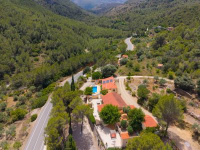 Vista aérea de la finca con dos viviendas, techos de teja y piscina azul, enclavada entre densos bosques verdes y una carretera sinuosa en un valle montañoso. Paneles solares visibles.
