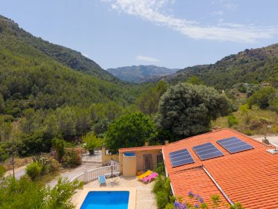 Vista aérea de la finca con tejado de teja, paneles solares y piscina azul, enmarcada por montañas boscosas y vegetación mediterránea.
