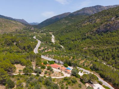 Vista aérea de la finca situada en un valle montañoso y boscoso, con piscina, paneles solares y edificios de tejado terracota, junto a una carretera serpenteante.