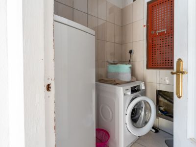 Zona de lavandería funcional con lavadora blanca, un congelador vertical, paredes alicatadas en beige y una distintiva rejilla de ventilación naranja. Tuberías expuestas son visibles.